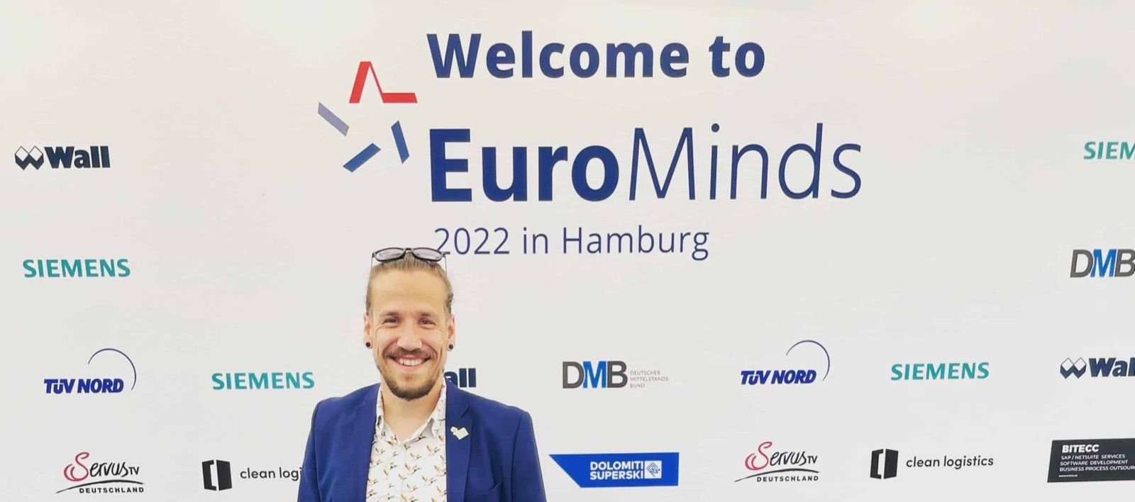 Carsten im Anzug vor der Begrüßungswand des EuroMinds-Gipfels 2022 in Hamburg.