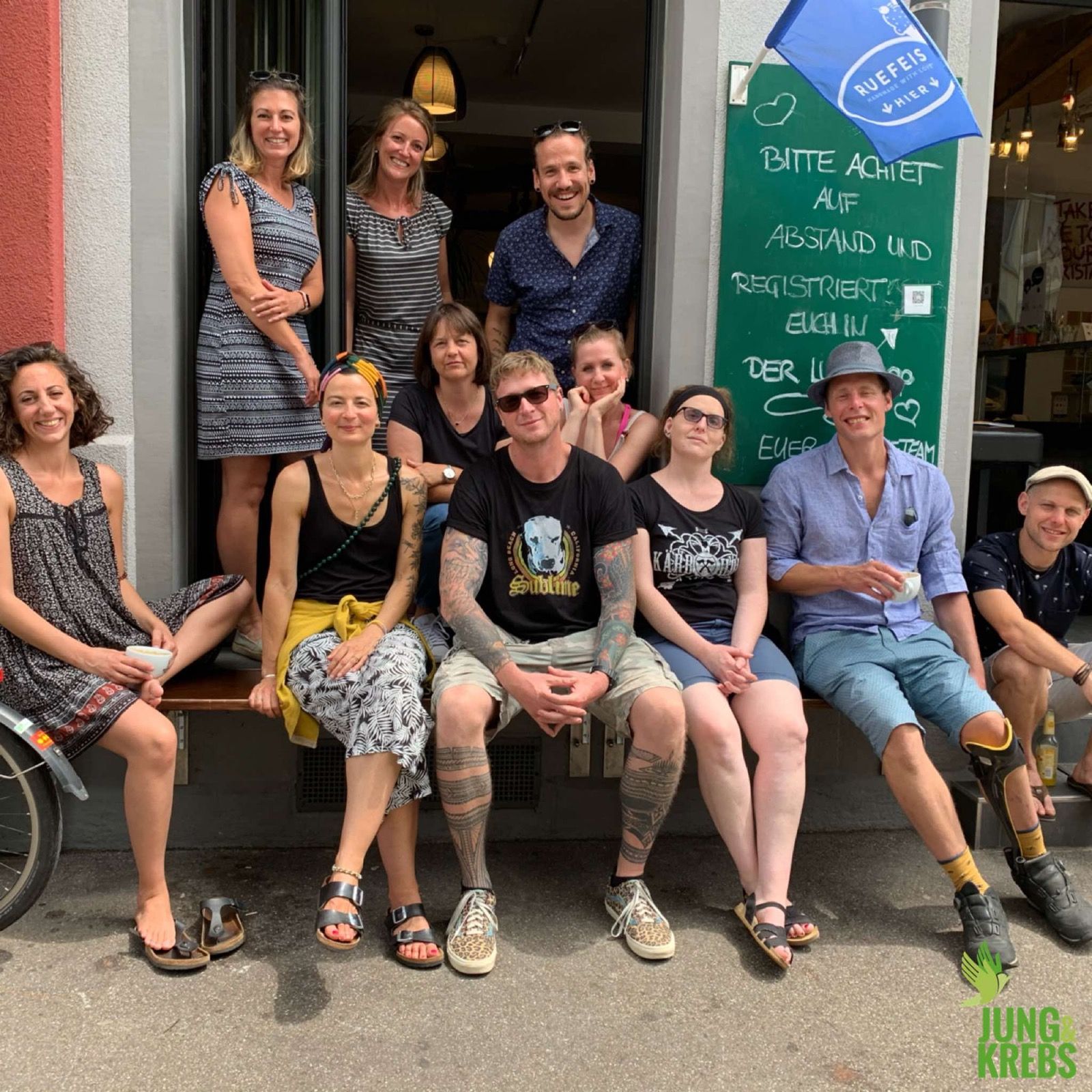 Rund fünfzehn Menschen sitzen und stehen gut gelaunt vor dem Eingang eines Cafés.