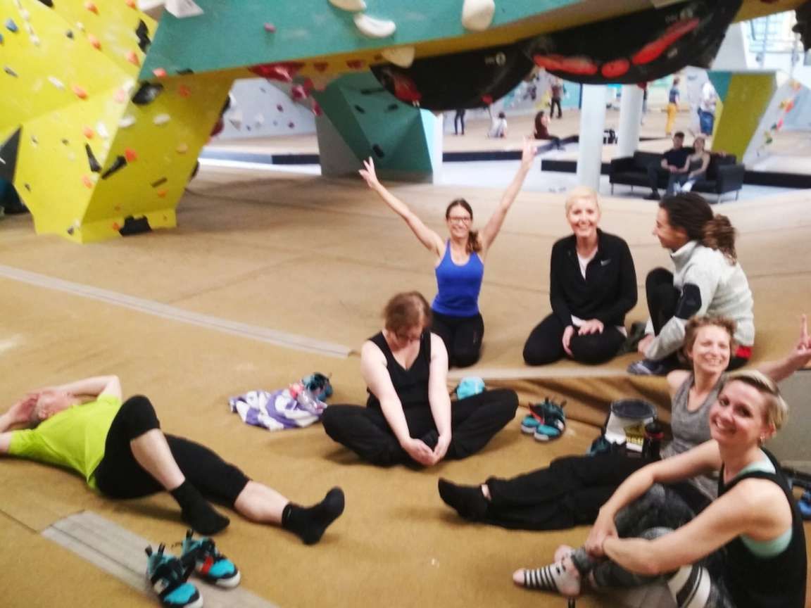 Eine Gruppe sitzt und liegt lachend auf der Matte einer Boulderhalle vor bunten Kletterwänden.
