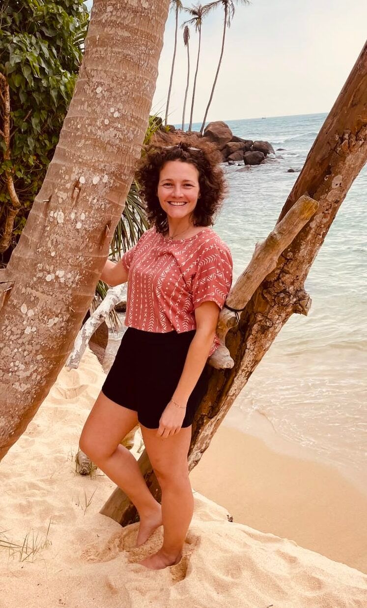 Desi lehnt lächelnd an einer Palme am Strand.