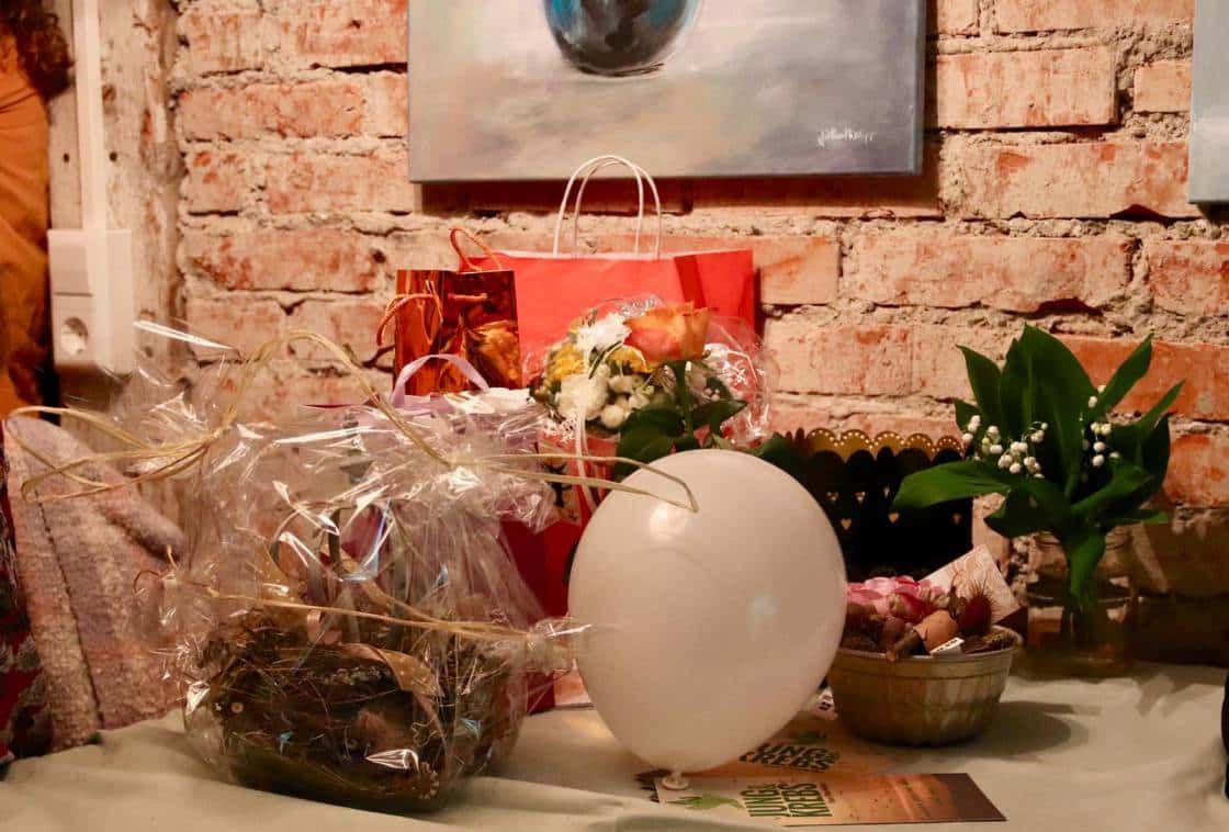 Ein Tisch vor einer Backsteinwand mit Blumen, einem Geschenkkorb, Geschenktüten und einem weißen Luftballon.