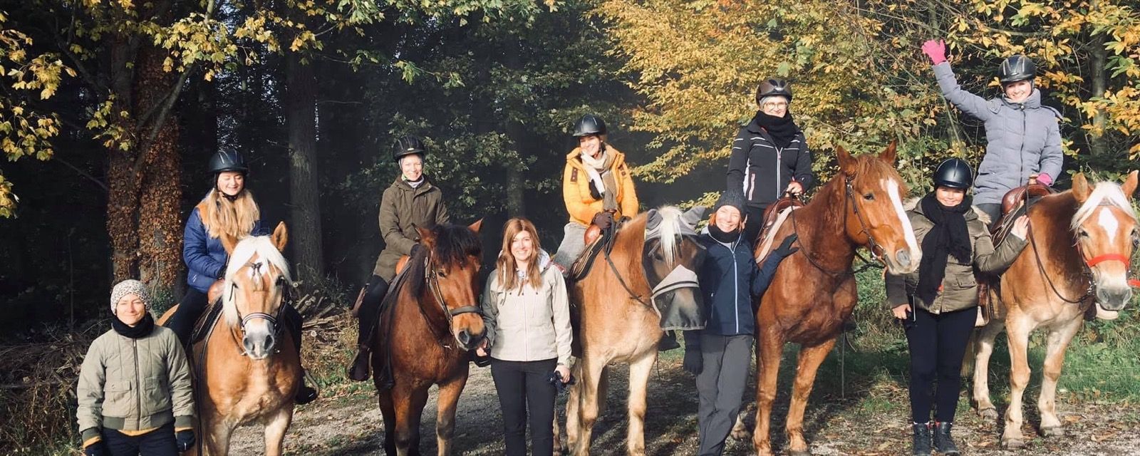 Mehrere Menschen zu Pferd und zu Fuß auf einem Waldweg im Herbstlicht.