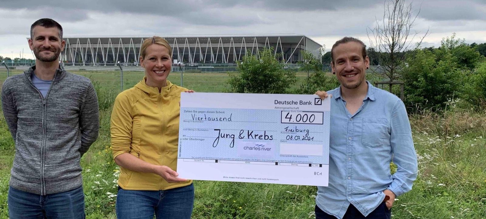 Zwei Männer und eine Frau halten auf einer Wiese einen Spendenscheck von Charles River über 4.000 Euro.