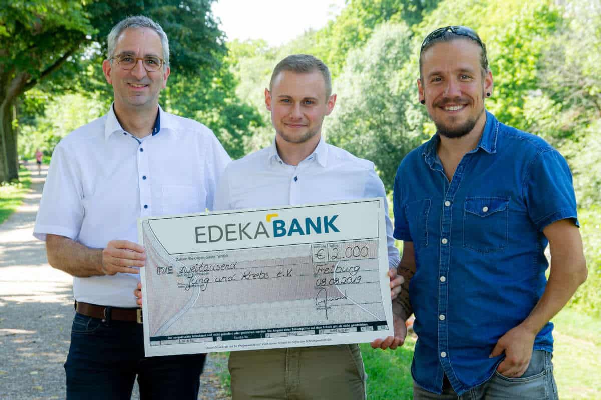 Drei Männer im Freien halten einen Spendenscheck der EDEKA BANK für Jung und Krebs e.V.