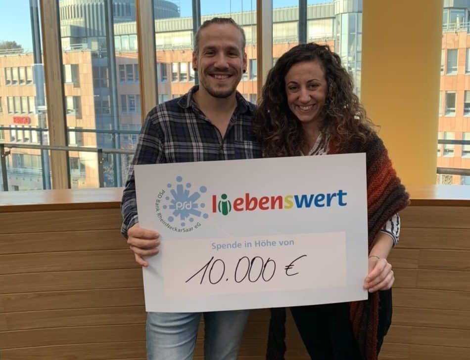 Ein Mann und eine Frau halten in einem Büro einen Scheck von liebenswert über 10.000 Euro.