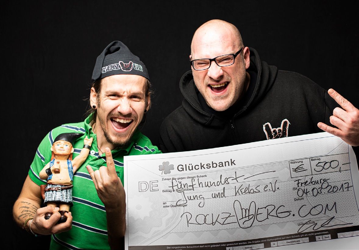 Zwei Männer lachen mit Rock-Geste in die Kamera und halten einen Spendenscheck über 500 Euro für Jung und Krebs e.V. von Rockzwerg.com.