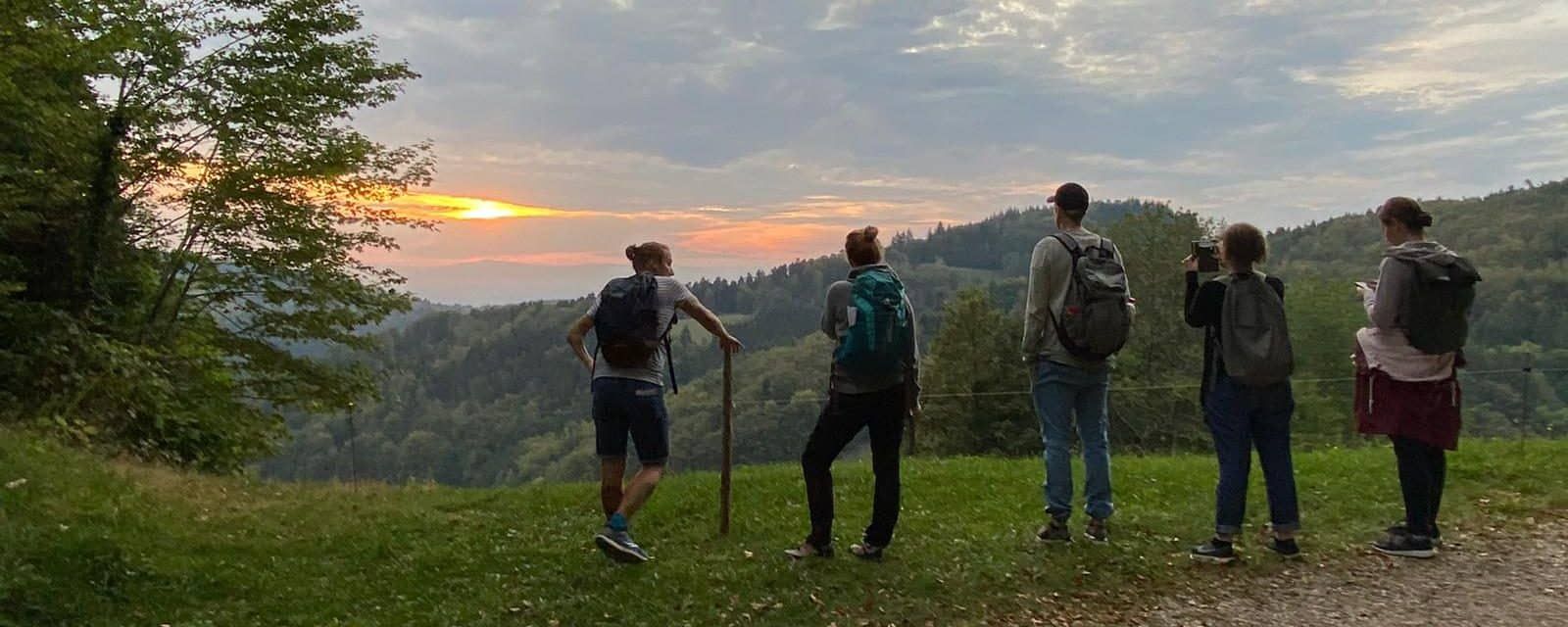 Fünf Menschen mit Rucksäcken stehen bei Sonnenuntergang auf einer Wiese und schauen über die Hügel des Schwarzwalds.