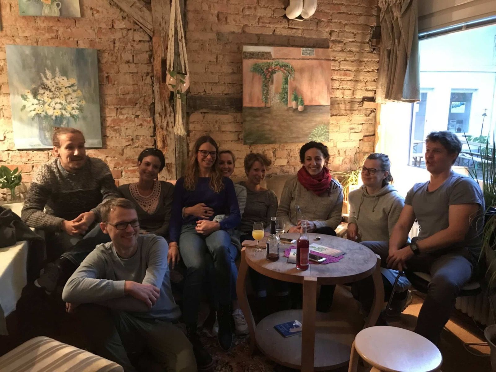 Gut ein Dutzend junge Menschen sitzt gemütlich um einen kleinen Tisch in einem Café mit Backsteinwand und unterhält sich.