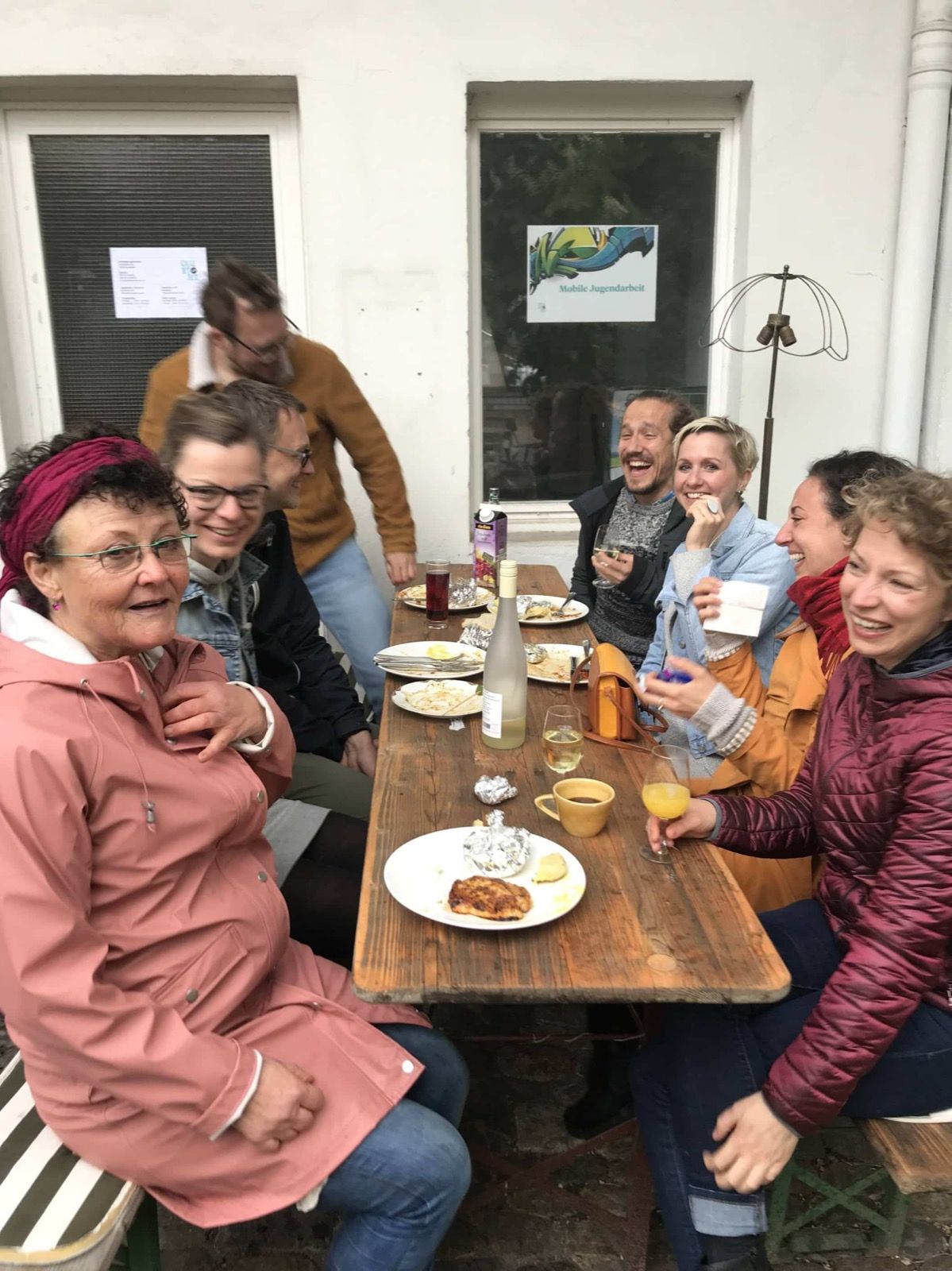Sieben Menschen sitzen lachend an einem langen Holztisch im Hof eines Cafés, auf dem Tisch Tassen und Kuchen.