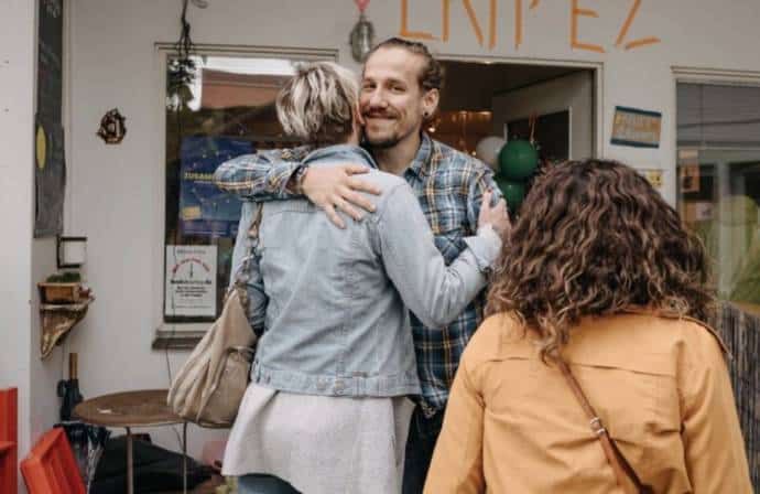 Zwei Menschen umarmen sich herzlich in der Tür eines Cafés, daneben eine junge Frau mit Locken.