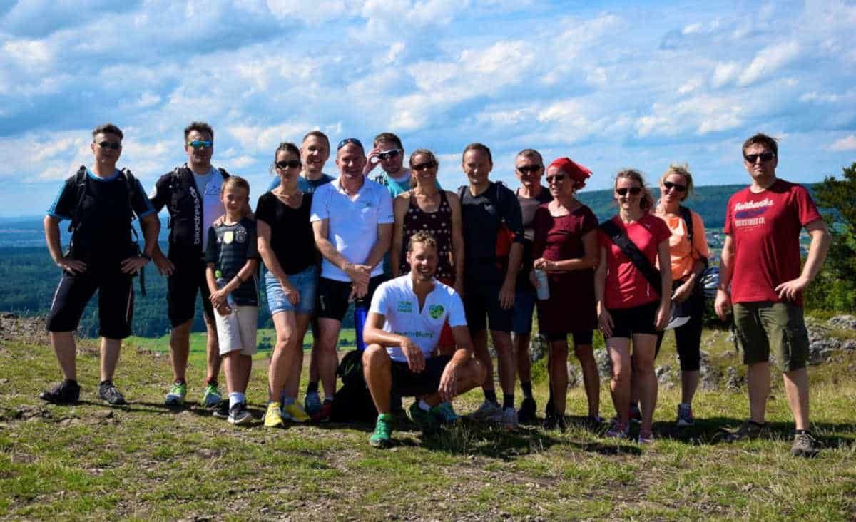 Eine Gruppe von gut fünfzehn Menschen in Wanderkleidung steht auf einer Wiese auf einem Hügel, dahinter Wald, Täler und blauer Himmel.