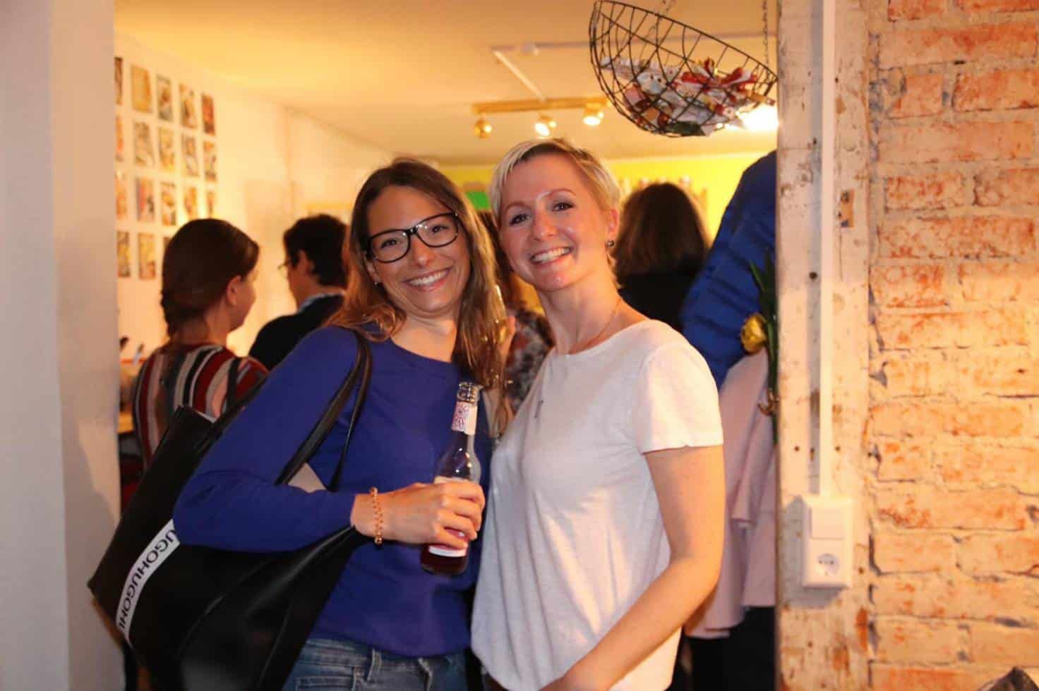 Zwei junge Frauen stehen Arm in Arm und lachen in die Kamera, im Hintergrund weitere Gäste vor einer Backsteinwand.